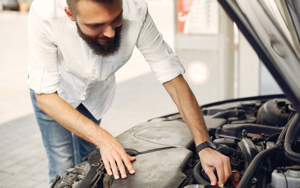 Hombre con barba revisando el motor abierto de un coche en una gasolinera.